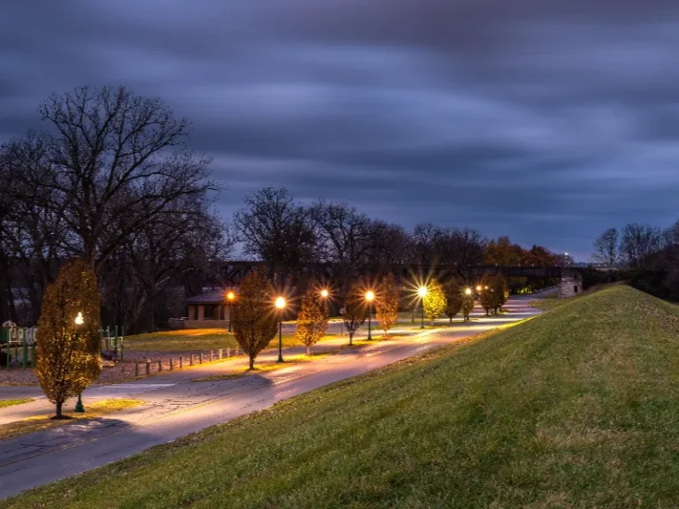 Streetlights illuminate a tree-lined path beside a grassy embankment under a cloudy evening sky.