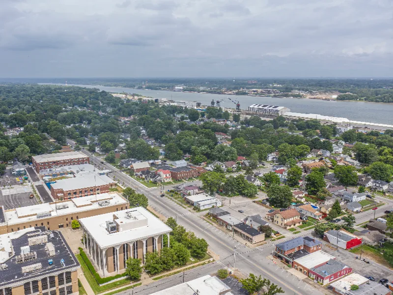 Aerial view of a suburban area with streets, houses, trees, and a river in the background under a cloudy sky.