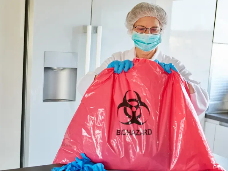 Person wearing protective gear holding a red biohazard waste bag in a kitchen setting.