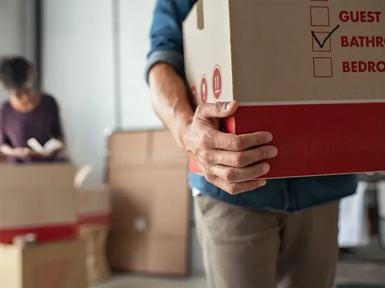 Person carrying a cardboard box labeled "Bathroom" with a woman reading in the background.