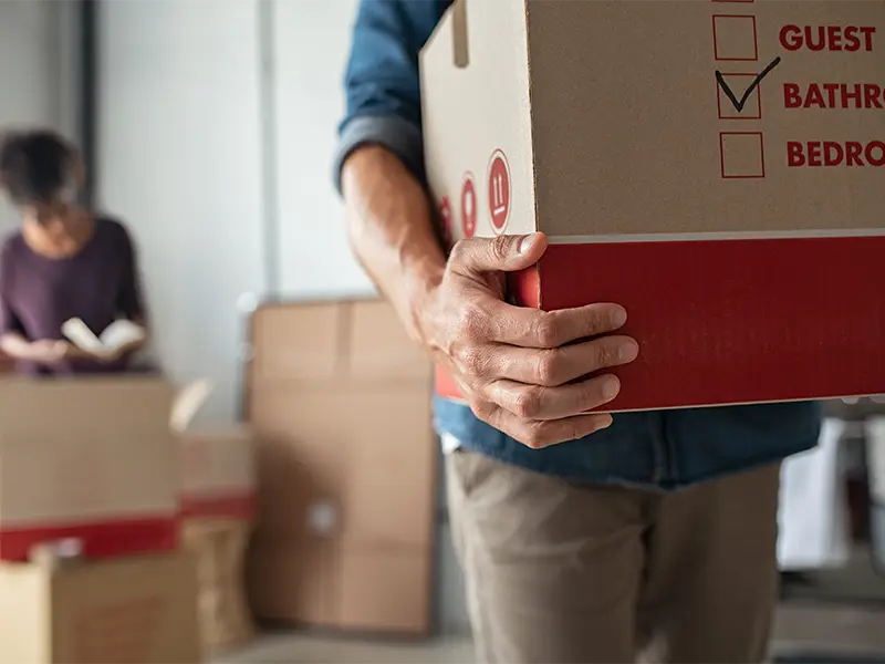 Person carrying a cardboard box labeled "Bathroom" with a woman reading in the background.