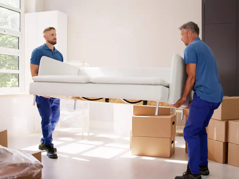 Two men in blue shirts carrying a white sofa in a bright room with cardboard boxes.