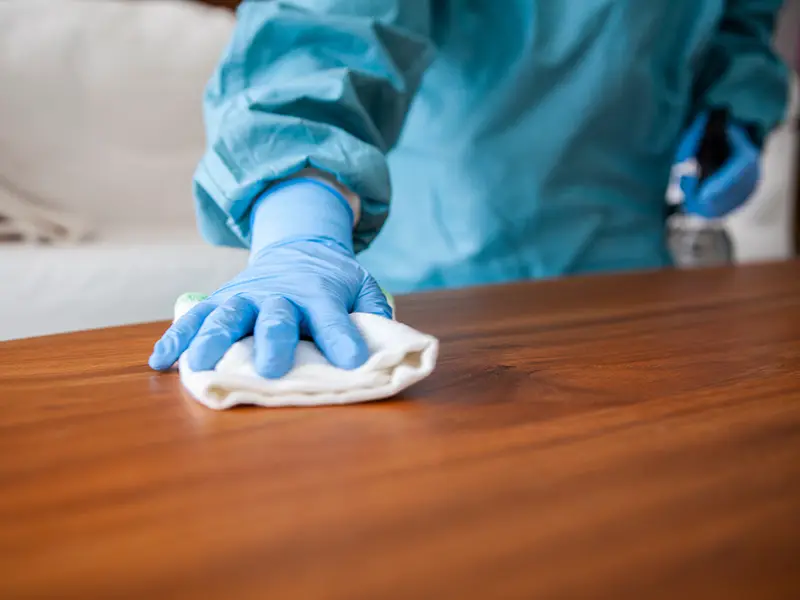 Hand in blue glove wiping a wooden surface with a white cloth.