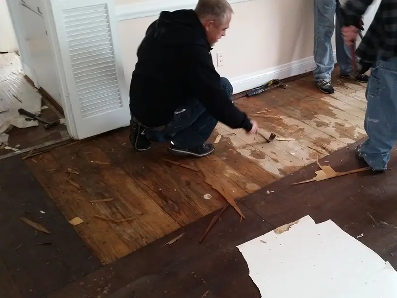 Man scraping damaged wooden floorboards in a room with two other people standing nearby.