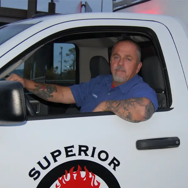 Man with tattoos wearing a blue shirt sitting in the driver's seat of a white truck with "Superior" logo.
