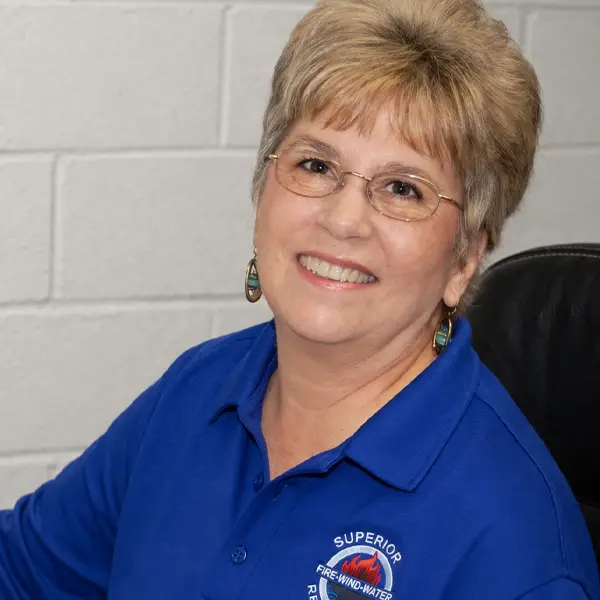 Smiling woman with glasses wearing a blue shirt with a fire rescue logo sitting in front of a gray brick wall.