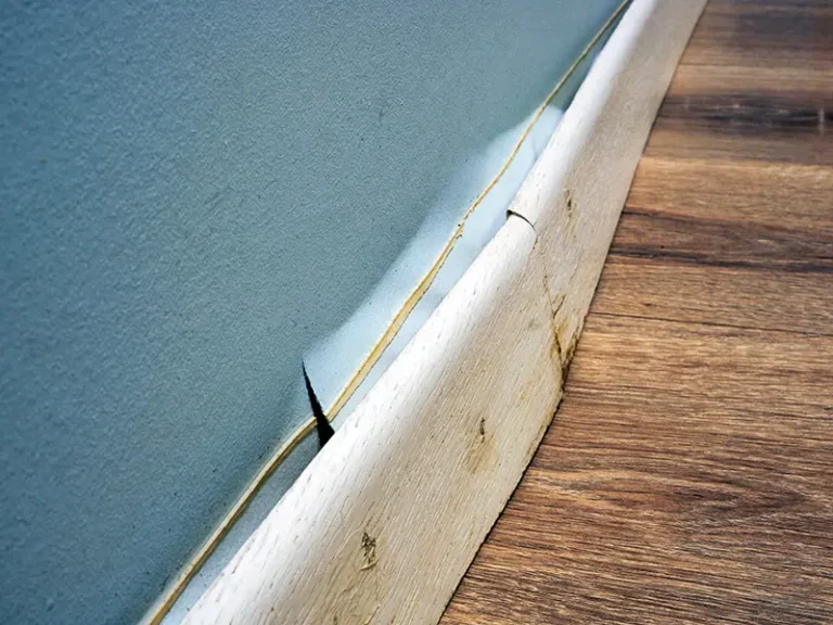 Peeling blue wall paint above a cracked white baseboard on a wooden floor.