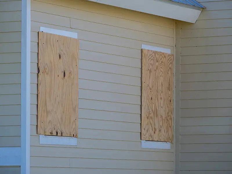 Two windows on a beige house are covered with plywood boards.